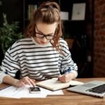 A woman sitting at her home office desk, using a calculator and writing in a notebook.