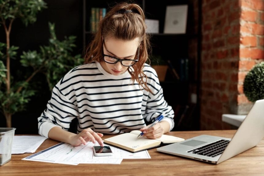 A woman sitting at her home office desk, using a calculator and writing in a notebook.